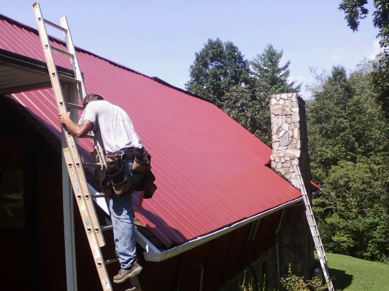 Roofer on a ladder beside a red metal roof and stone chimney — Soffit & Fascia Repair by Roofing Brothers in Atalissa
